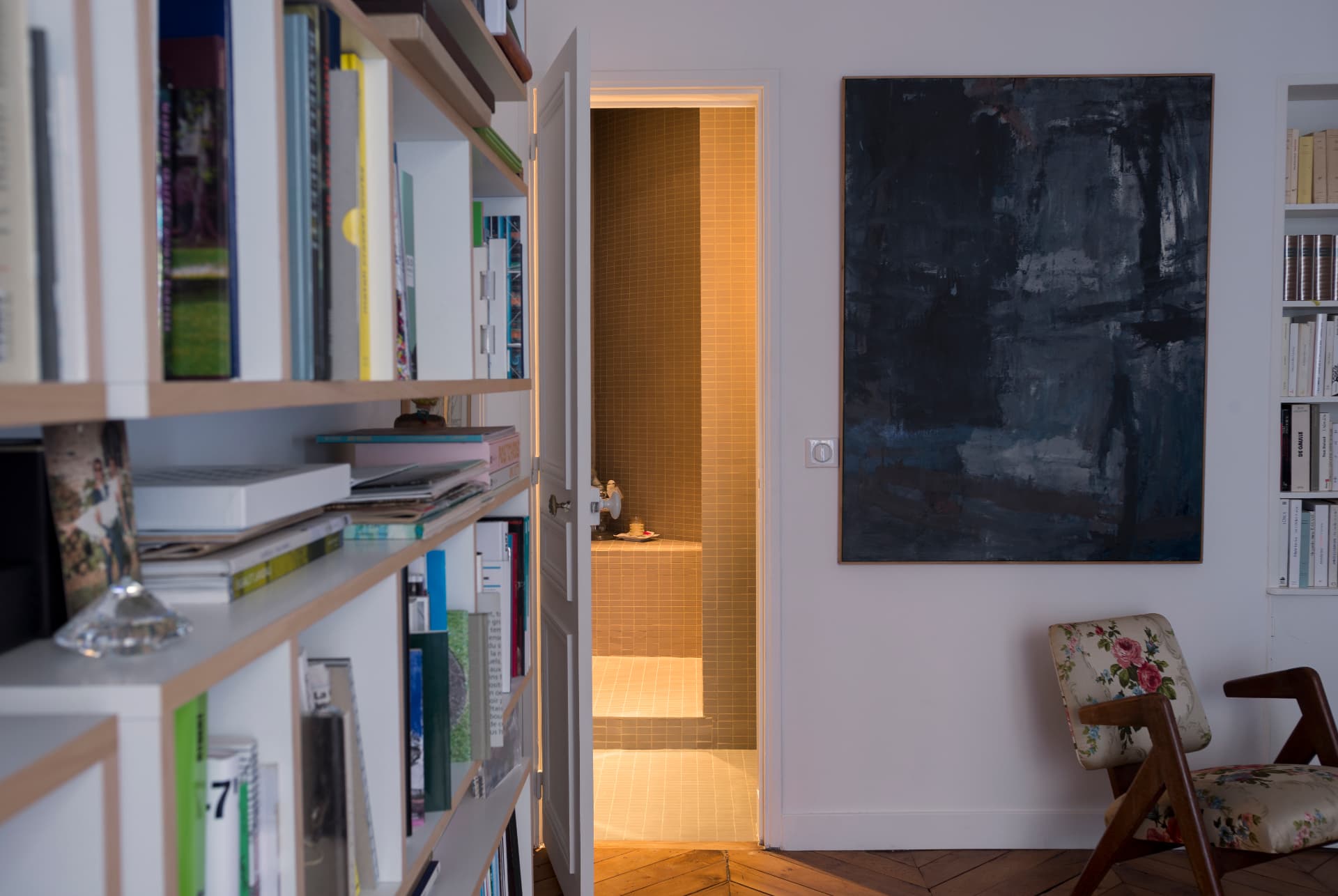 Library wall of books beside a large abstract painting and a lit bathroom beyond