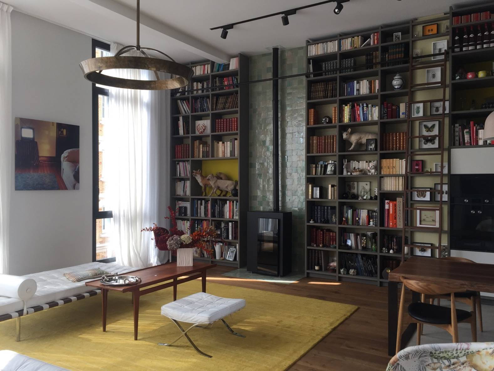 Library wall with wood stove, zellige tile chimney and walnut ladder above a yellow rug