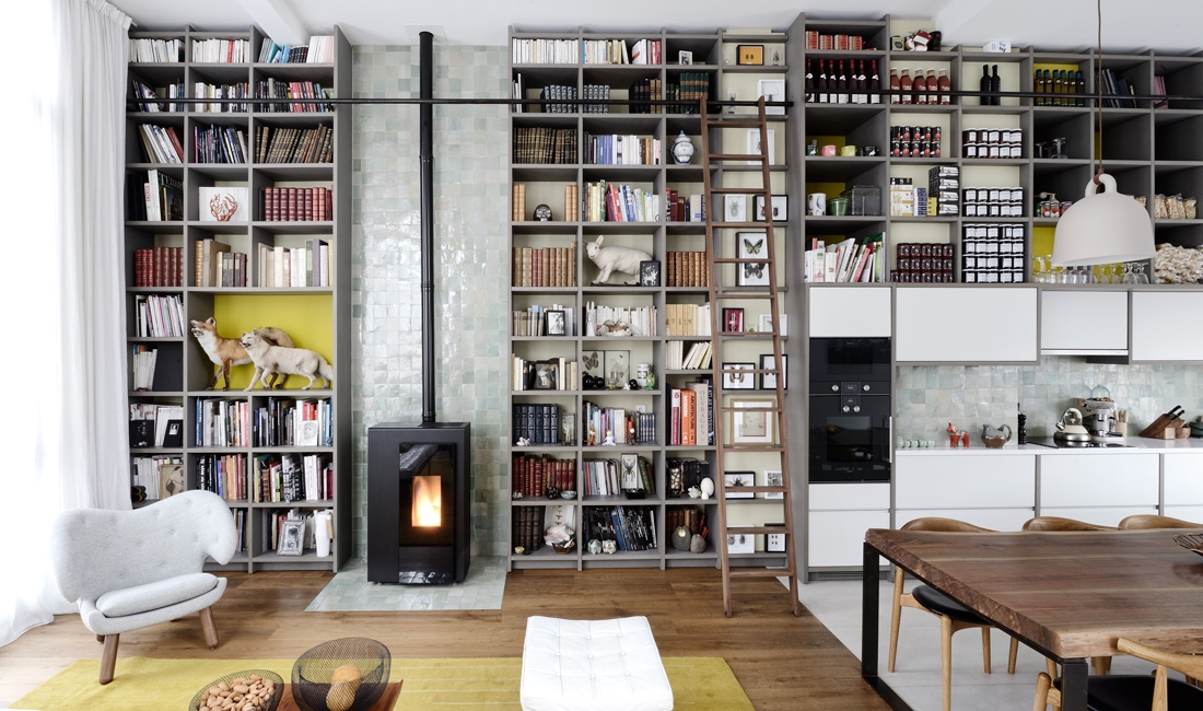 Cathedral-height living room with white armchair, yellow rug and wood stove backed by zellige tile