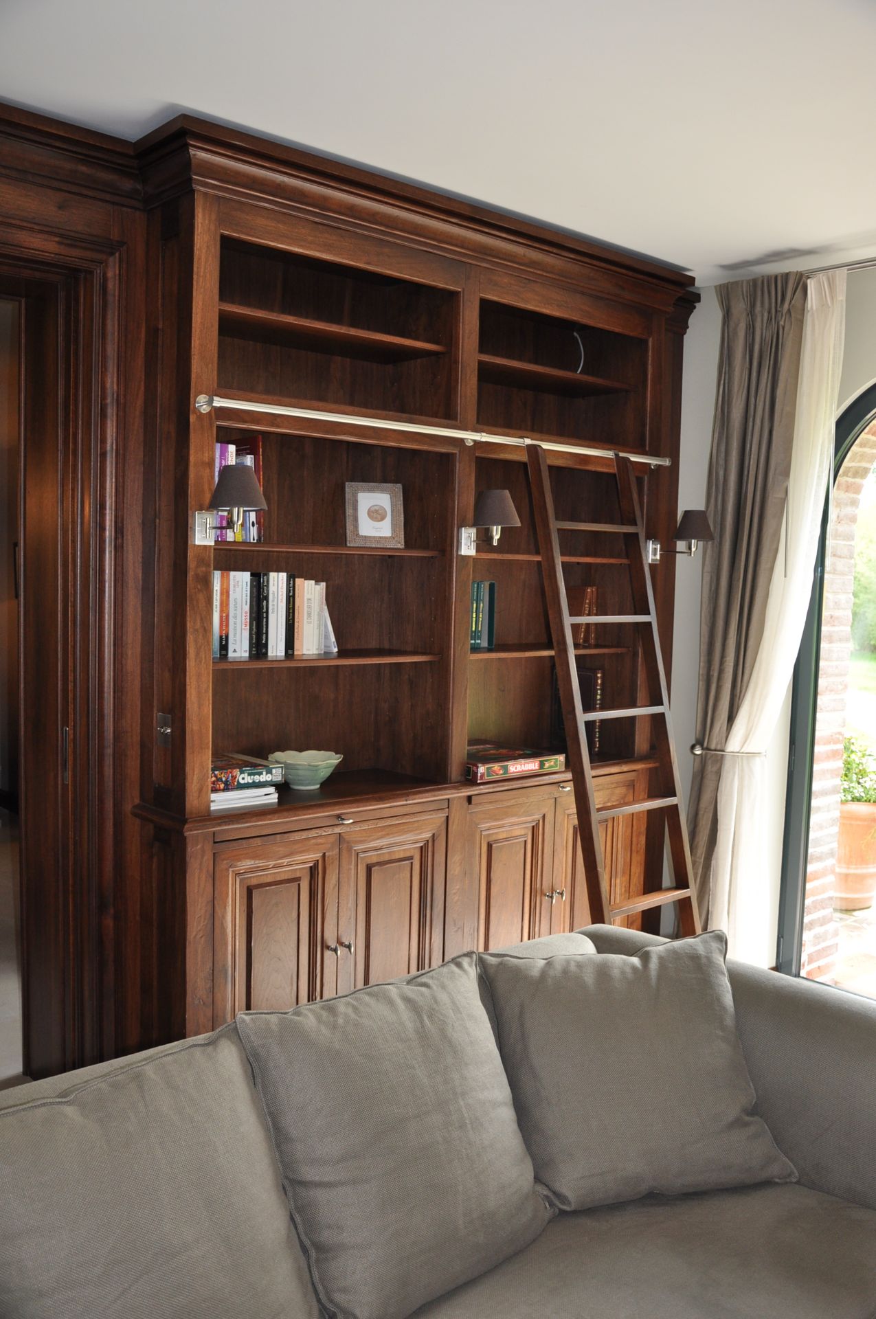 Walnut library bookcase with rolling ladder and reading sconces beside a linen sofa